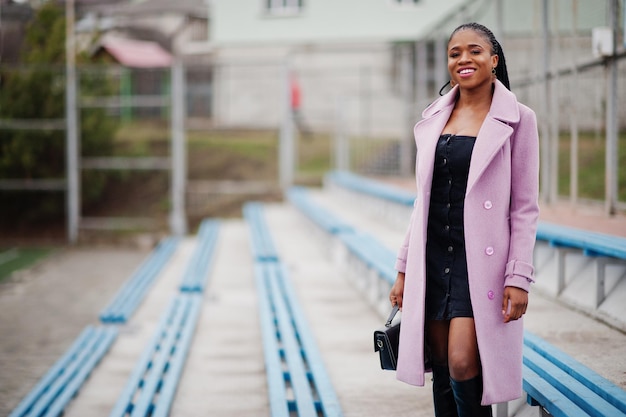 Junge stilvolle schöne afrikanisch-amerikanische Frau auf der Straße an den Tribünen des Stadions, die Mode-Outfit-Mantel-Handtasche trägt
