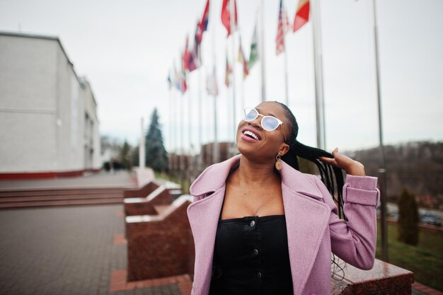 Junge stilvolle schöne afrikanisch-amerikanische Frau auf der Straße mit Mode-Outfit-Mantel und Brille gegen Flaggen verschiedener Länder der Welt