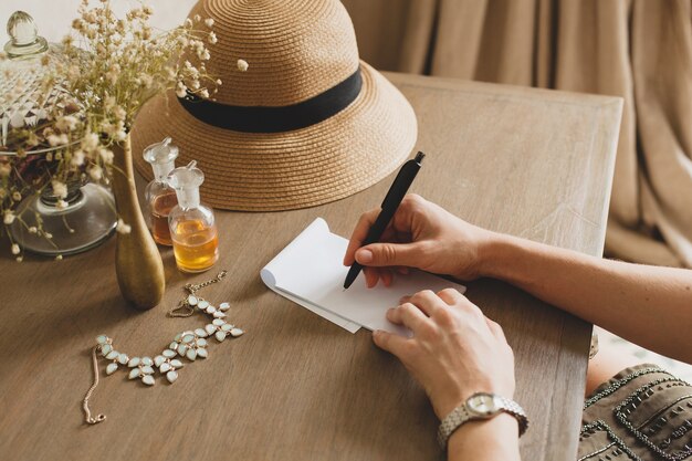 Junge stilvolle schöne Frau, die am Tisch im Hotelzimmer des Resorts sitzt und einen Brief schreibt, Stift, Strohhut, Vintage-Stil, Hände Nahaufnahme, Details, Zubehör, Reisetagebuch hält