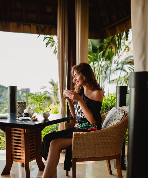 Junge stilvolle schöne Frau, die am tropischen Resortcafé sitzt