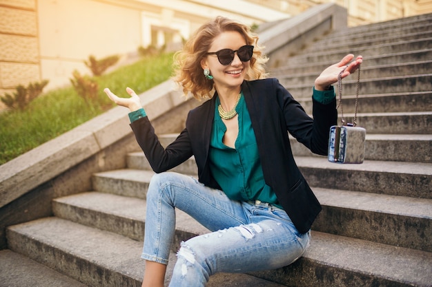 Junge stilvolle schöne Frau, die auf Treppe in der Stadtstraße sitzt und Jeans, schwarze Jacke, grüne Bluse, Sonnenbrille, Geldbeutel, eleganten Stil, Sommermodetrend, lächelnd trägt