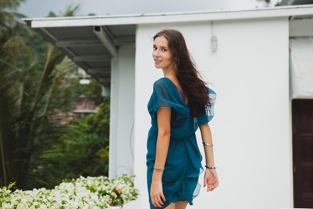 Junge stilvolle schöne Frau im blauen Kleid, Sommermodetrend, Urlaub, Garten, tropische Hotelterrasse, lächelnd, gehend