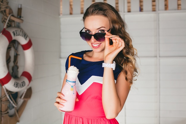 Junge stilvolle schöne Frau im Seecafé, Trinkcocktail Smoothie, Sonnenbrille, Flirty, Resort-Stil, modisches Outfit, Lächeln, Marinefarben Kleid, Anker und Rettungsring auf Hintergrund, schockiert