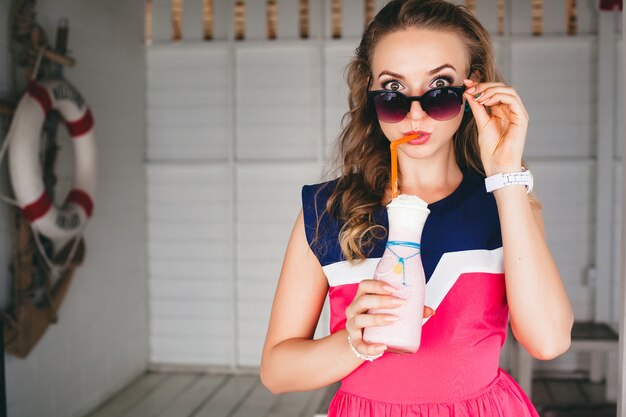 Junge stilvolle schöne Frau im Seecafé, Trinkcocktail Smoothie, Sonnenbrille, Flirty, Resort-Stil, modisches Outfit, Lächeln, Marinefarben Kleid, Anker und Rettungsring auf Hintergrund, schockiert