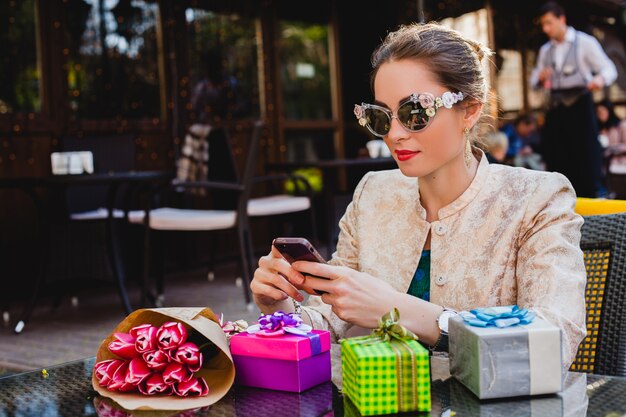 Junge stilvolle schöne Frau in der Mode-Sonnenbrille, die am Café sitzt und Telefon hält