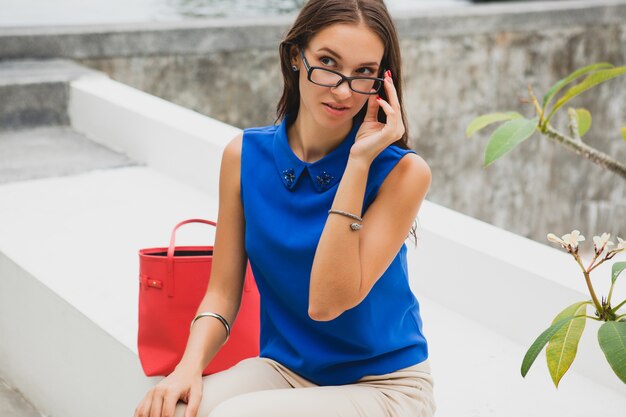 Junge stilvolle schöne Frau, Sommermodetrend, blaue Bluse, rote Tasche, Brille, tropisches Villenresort, Urlaub, flirty