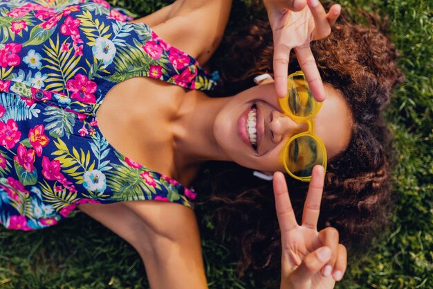 Junge stilvolle schwarze Frau, die Musik auf kabellosen Kopfhörern hört, die Spaß im Park, Sommermodeart, buntes Hipster-Outfit haben, auf Gras liegend, Ansicht von oben