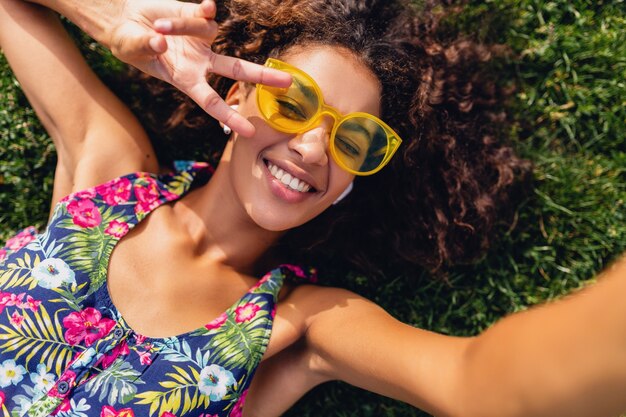 Junge stilvolle schwarze Frau, die Musik auf kabellosen Kopfhörern hört, die Spaß im Park, Sommermodeart, buntes Hipster-Outfit haben, auf Gras liegend, Ansicht von oben