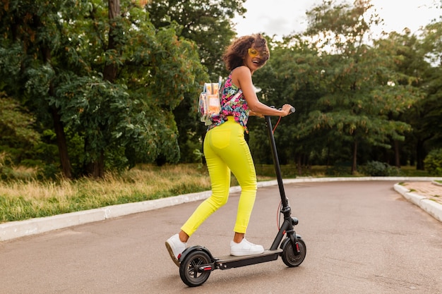Junge stilvolle schwarze Frau, die Spaß im Park reitet auf elektrischem Tretroller im Sommermode-Stil, buntes Hipster-Outfit, Rucksack und gelbe Sonnenbrille tragend