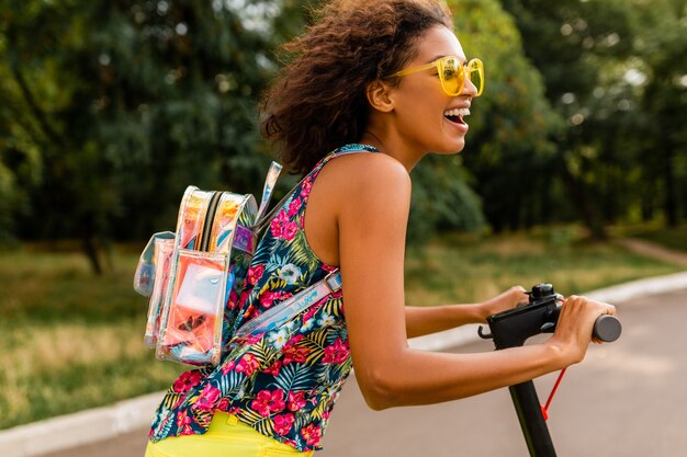 Junge stilvolle schwarze Frau, die Spaß im Park reitet auf elektrischem Tretroller im Sommermode-Stil, buntes Hipster-Outfit, Rucksack und gelbe Sonnenbrille tragend