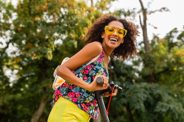 Junge stilvolle schwarze Frau, die Spaß im Park reitet auf elektrischem Tretroller im Sommermode-Stil, buntes Hipster-Outfit, Rucksack und gelbe Sonnenbrille tragend