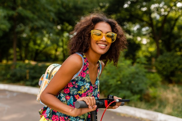 Junge stilvolle schwarze Frau, die Spaß im Park reitet auf elektrischem Tretroller im Sommermode-Stil, buntes Hipster-Outfit, Rucksack und gelbe Sonnenbrille tragend