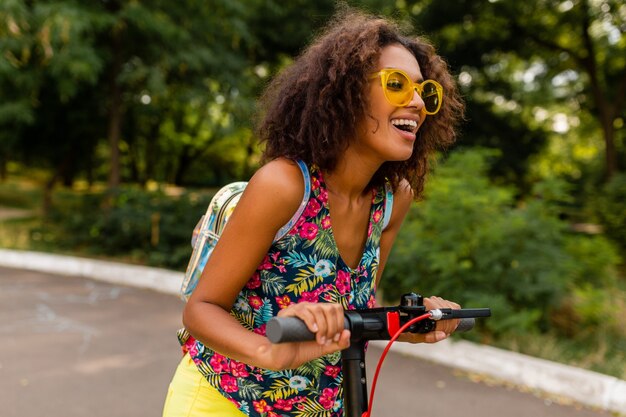 Junge stilvolle schwarze Frau, die Spaß im Park reitet auf elektrischem Tretroller im Sommermode-Stil, buntes Hipster-Outfit, Rucksack und gelbe Sonnenbrille tragend