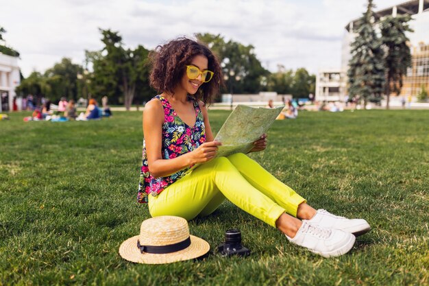 Junge stilvolle schwarze Frau, die Spaß im Park-Sommer-Modestil, buntes Hipster-Outfit, auf Gras sitzend, Reisender mit einer Karte und Strohhut hat