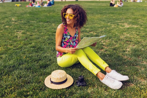 Junge stilvolle schwarze Frau, die Spaß im Park-Sommermodenstil, buntes Hipster-Outfit hat, auf Gras sitzend, Reisender mit einer Karte