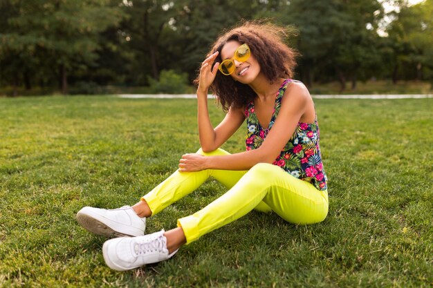 Junge stilvolle schwarze Frau, die Spaß im Park-Sommermodenstil, buntes Hipster-Outfit hat, das auf Gras sitzt