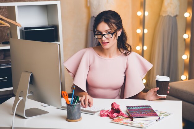 Junge stilvolle sexy Frau im rosa Luxuskleid, Sommertrend, schicker Stil, Modedesigner, der im Büro am Computer arbeitet