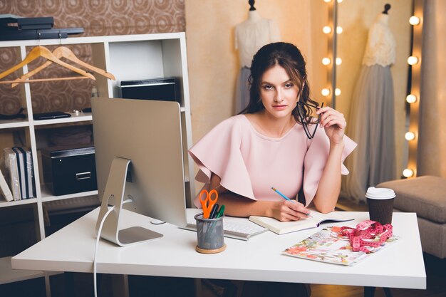 Junge stilvolle sexy Frau im rosa Luxuskleid, Sommertrend, schicker Stil, Modedesigner, der im Büro am Computer arbeitet