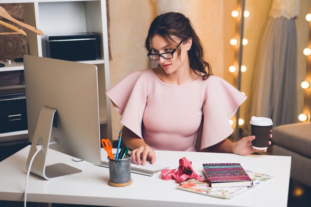 Junge stilvolle sexy Frau im rosa Luxuskleid, Sommertrend, schicker Stil, Modedesigner, der im Büro am Computer arbeitet