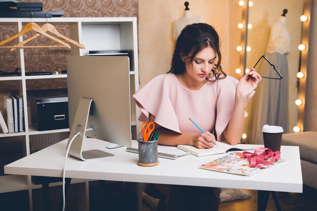 Junge stilvolle sexy Frau im rosa Luxuskleid, Sommertrend, schicker Stil, Modedesigner, der im Büro am Computer arbeitet