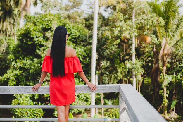 Junge stilvolle sexy Frau im roten Sommerkleid stehend auf Terrasse im tropischen Hotel, Palmenhintergrund, langes schwarzes Haar, Sonnenbrille, ethnische Ohrringe, Sonnenbrille, die sich freuen