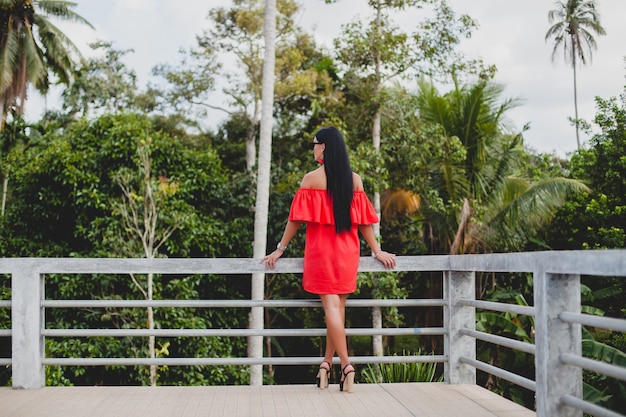 Junge stilvolle sexy Frau im roten Sommerkleid stehend auf Terrasse im tropischen Hotel, Palmenhintergrund, langes schwarzes Haar, Sonnenbrille, ethnische Ohrringe, Sonnenbrille, die sich freuen