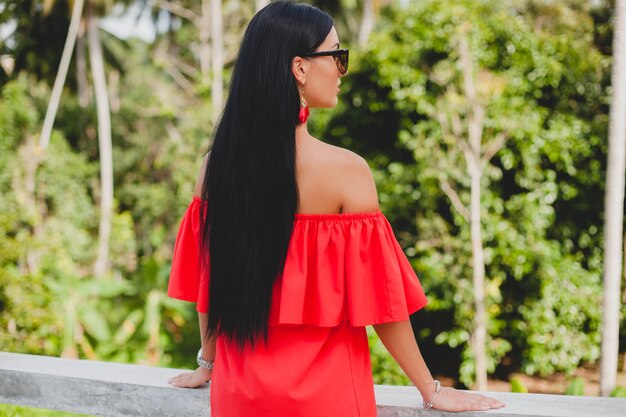 Junge stilvolle sexy Frau im roten Sommerkleid stehend auf Terrasse im tropischen Hotel, Palmenhintergrund, langes schwarzes Haar, Sonnenbrille, ethnische Ohrringe, Sonnenbrille, die sich freuen