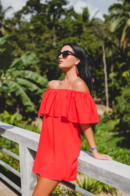Junge stilvolle sexy Frau im roten Sommerkleid stehend auf Terrasse im tropischen Hotel, Palmenhintergrund, langes schwarzes Haar, Sonnenbrille, ethnische Ohrringe, Sonnenbrille, lächelnd