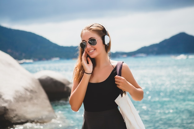 Junge stilvolle sexy Hipster-Frau mit einer Einkaufstasche während des Urlaubs, Fliegersonnenbrille, Kopfhörer, Musik hören, glücklich, Sonne genießen, tropische Insel blaue Lagunenlandschaft