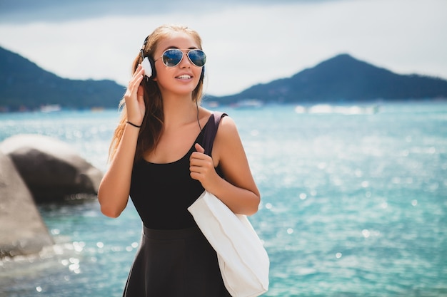 Junge stilvolle sexy Hipster-Frau mit einer Einkaufstasche während des Urlaubs, Fliegersonnenbrille, Kopfhörer, Musik hören, glücklich, Sonne genießen, tropische Insel blaue Lagunenlandschaft