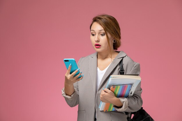 Junge Studentin der Vorderansicht im grauen Mantel, der das Halten von Büchern unter Verwendung eines Telefons auf dem Universitätshochschulstudium des rosa Hintergrundunterrichts aufwirft