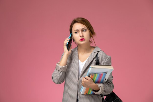 Junge Studentin der Vorderansicht im grauen Mantel, der hält Bücher hält, die am Telefon auf dem rosa Hintergrundlektionsuniversitätshochschulstudienbuch sprechen