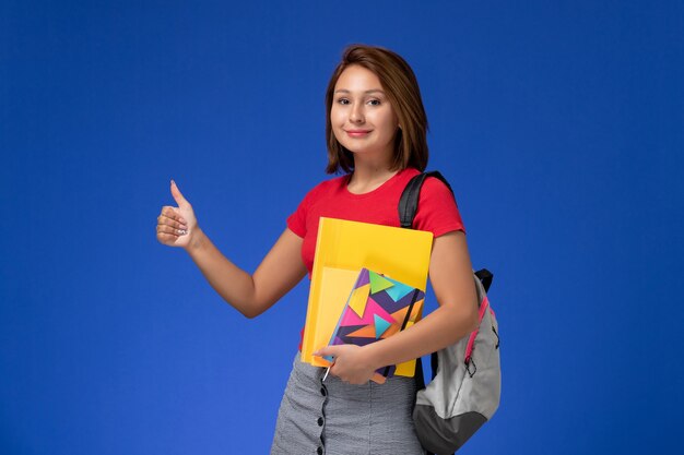 Junge Studentin der Vorderansicht im roten Hemd, das Rucksack hält, der Dateien und Heft auf dem blauen Hintergrund hält.