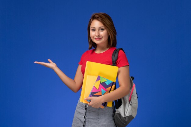 Junge Studentin der Vorderansicht im roten Hemd, das Rucksack hält, der Dateien und Heft auf dem blauen Hintergrund hält.