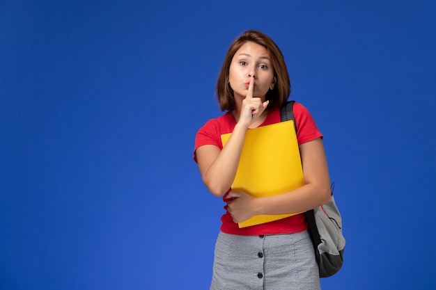 Junge Studentin der Vorderansicht im roten Hemd mit dem Rucksack, der gelbe Dateien hält, zeigen Stillezeichen auf hellblauem Hintergrund.
