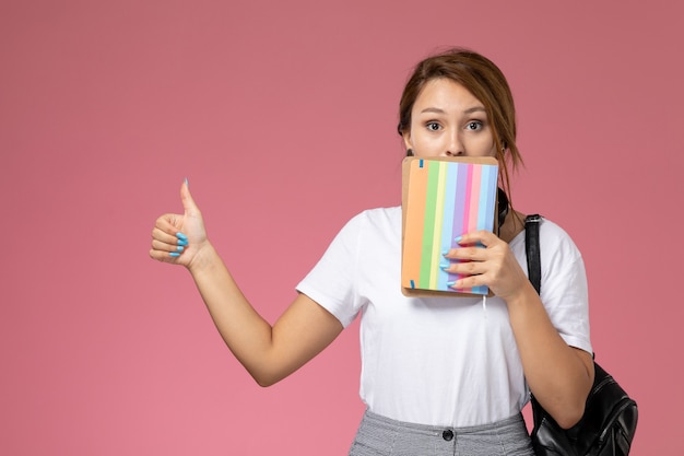 Junge Studentin der Vorderansicht im weißen T-Shirt und in der grauen Hose mit Heft in ihren Händen auf den Universitätshochschulen des rosa Hintergrundunterrichts