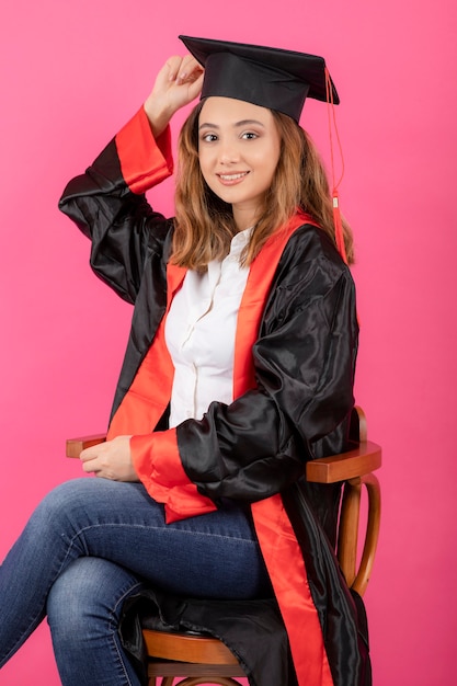 Kostenloses Foto junge studentin, die ein abschlusskleid trägt und auf einem stuhl an einer rosa wand sitzt.