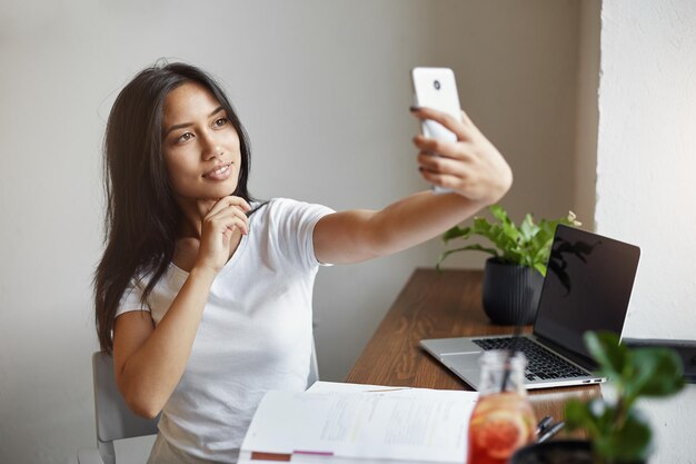 Junge Studentin, die während der Sommerpause ein Selfie im Café macht und sich darüber lustig macht, wie sie am Laptop arbeitet und von ihrem eigenen Online-Geschäft träumt