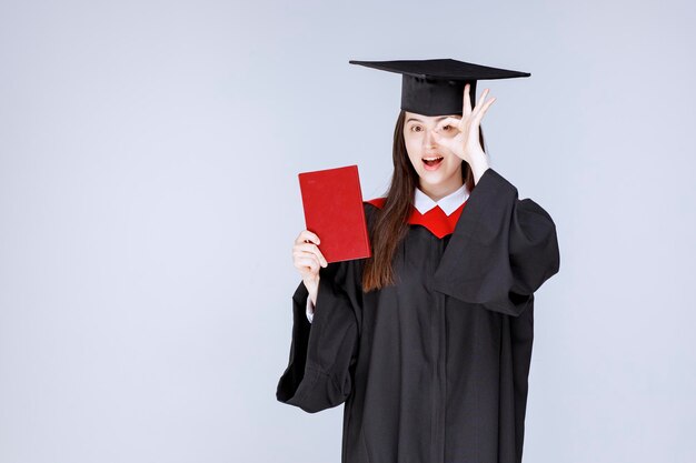 Junge Studentin im akademischen Gewand mit Buch, das OK-Zeichen zeigt. Foto in hoher Qualität