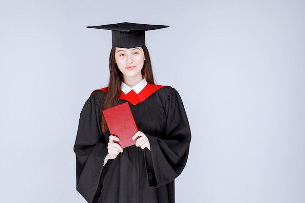 Junge Studentin im akademischen Kleid, die rotes Buch zur Kamera zeigt. Foto in hoher Qualität