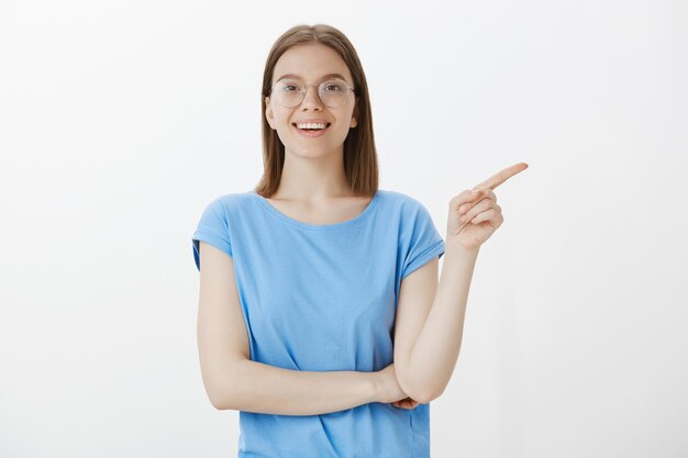 Junge Studentin in Gläsern, die lächeln und Finger rechts zeigen, fasziniert schauen