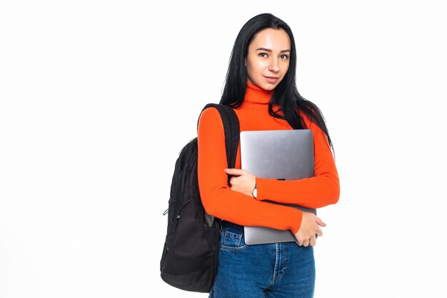 Junge Studentin isoliert auf grauer Wand, lächelt in die Kamera, drückt Laptop auf die Brust, trägt Rucksack, bereit, zum Studium zu gehen, neues Projekt zu starten und neue Ideen vorzuschlagen.