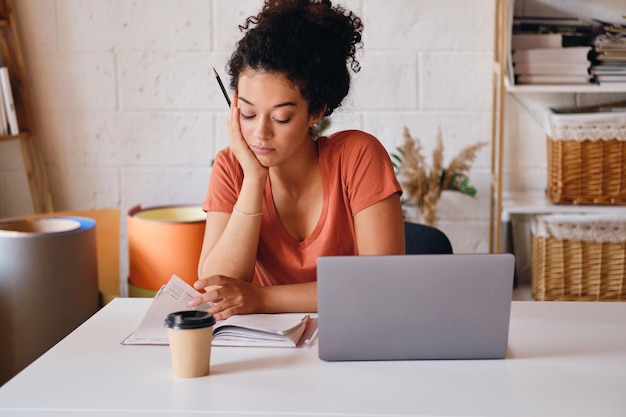 Junge Studentin mit dunklem, lockigem Haar, die mit Laptop und einer Tasse Kaffee am Tisch sitzt, um sich an die Hand zu lehnen und nachdenklich in einem gemütlichen Zuhause zu studieren