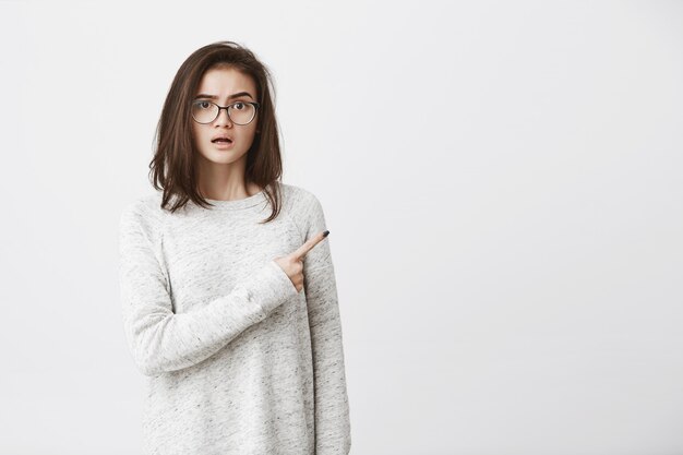 junge süße Frau, die Brille und weißes Sweatshirt trägt, zeigt mit verwundertem und verwirrtem Ausdruck zur Seite.