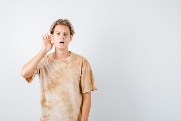 Junge Teenager im T-Shirt, die Hand hinter dem Ohr halten und verwirrt aussehen, Vorderansicht.