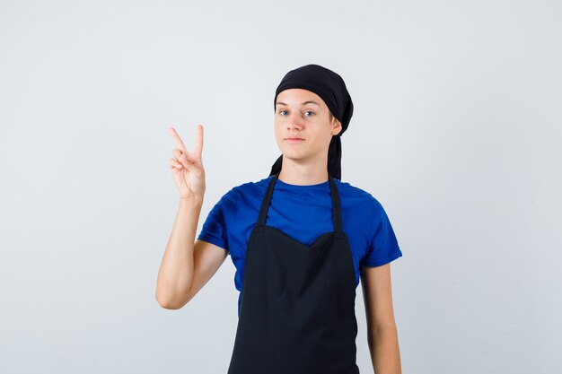 Junge Teenager-Koch im T-Shirt, Schürze mit V-Zeichen und stolz aussehend, Vorderansicht.