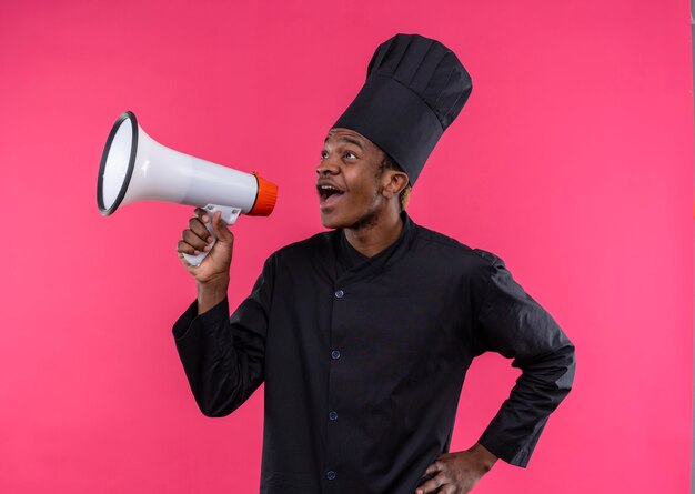 Junge überraschte afroamerikanische Köchin in der Kochuniform hält Lautsprecher lokalisiert auf rosa Wand