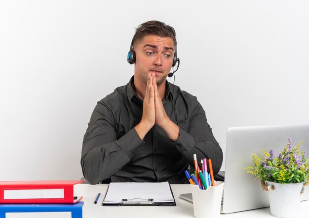 Junge überraschte blonde Büroangestellte Mann auf Kopfhörern sitzt am Schreibtisch mit Bürowerkzeugen, die Laptop betrachten, hält Hände zusammen lokalisiert auf weißem Hintergrund mit Kopienraum