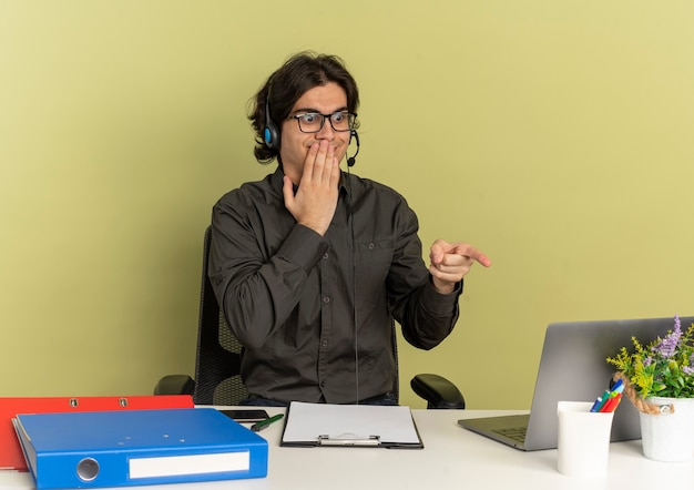 Kostenloses Foto junge überraschte büroangestellte mann auf kopfhörern in optischen gläsern sitzt am schreibtisch mit bürowerkzeugen, die auf laptop lokalisiert auf grünem hintergrund mit kopienraum suchen und zeigen