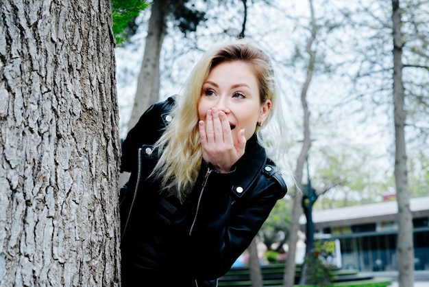 Kostenloses Foto junge überraschte frau, die sich hinter dem baum in einem park versteckt, der schwarze lederjacke trägt